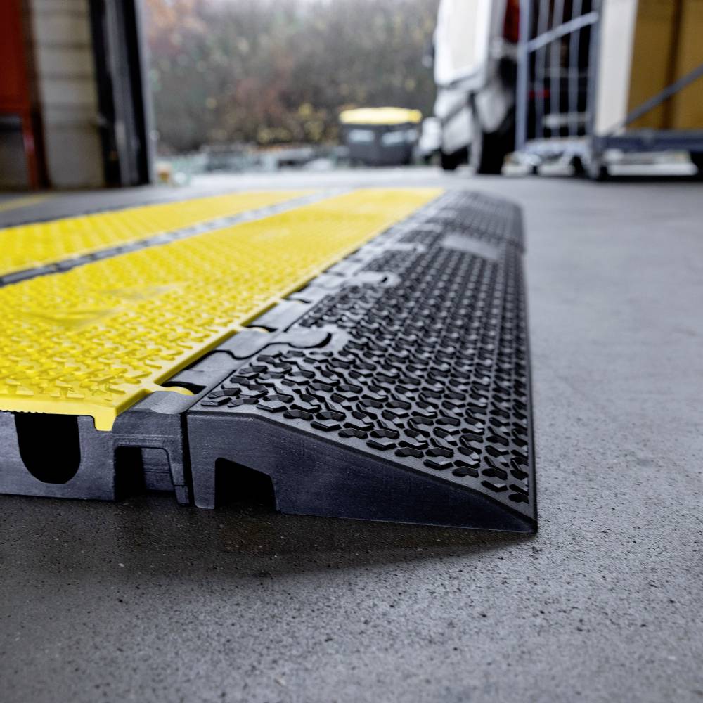 Eine flache schwarze und gelbe Kabelbrücke auf einem Betonboden in einer Garage, teilweise von einem Fahrzeug im Hintergrund verdeckt.