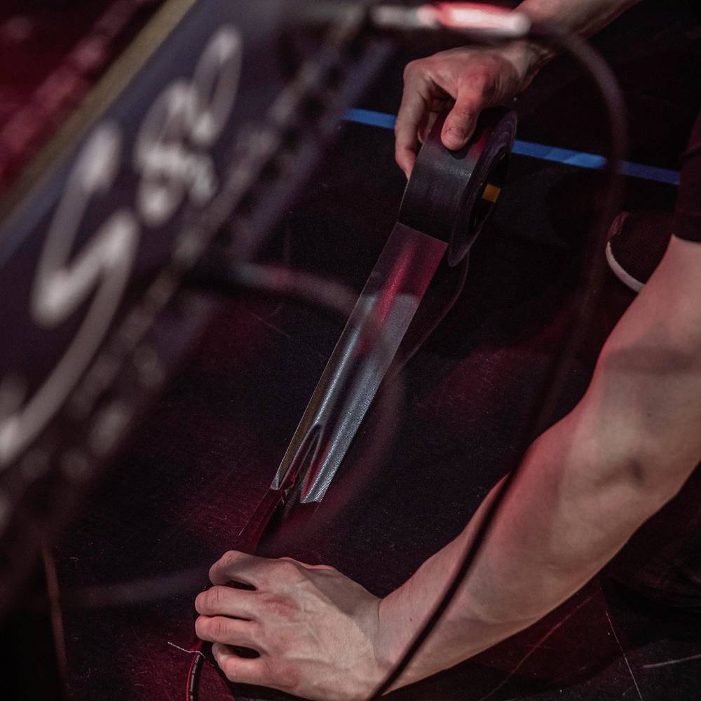 Eine Person schneidet mit einem Messer schwarzes Klebeband auf einer dunklen Oberfläche. Die Umgebung ist dunkel und unscharf.