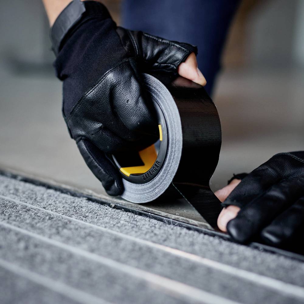 Eine Person mit schwarzen Handschuhen zieht einen Streifen schwarzes Klebeband von einer Rolle ab und klebt es auf eine Oberfläche.