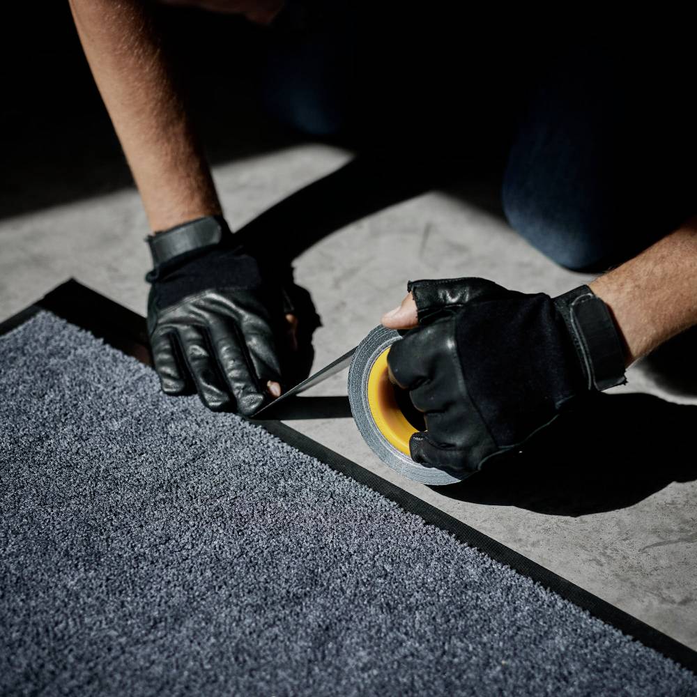 Eine Person mit Handschuhen schneidet mit einem Cuttermesser einen Teppich auf einer grauen Bodenoberfläche.