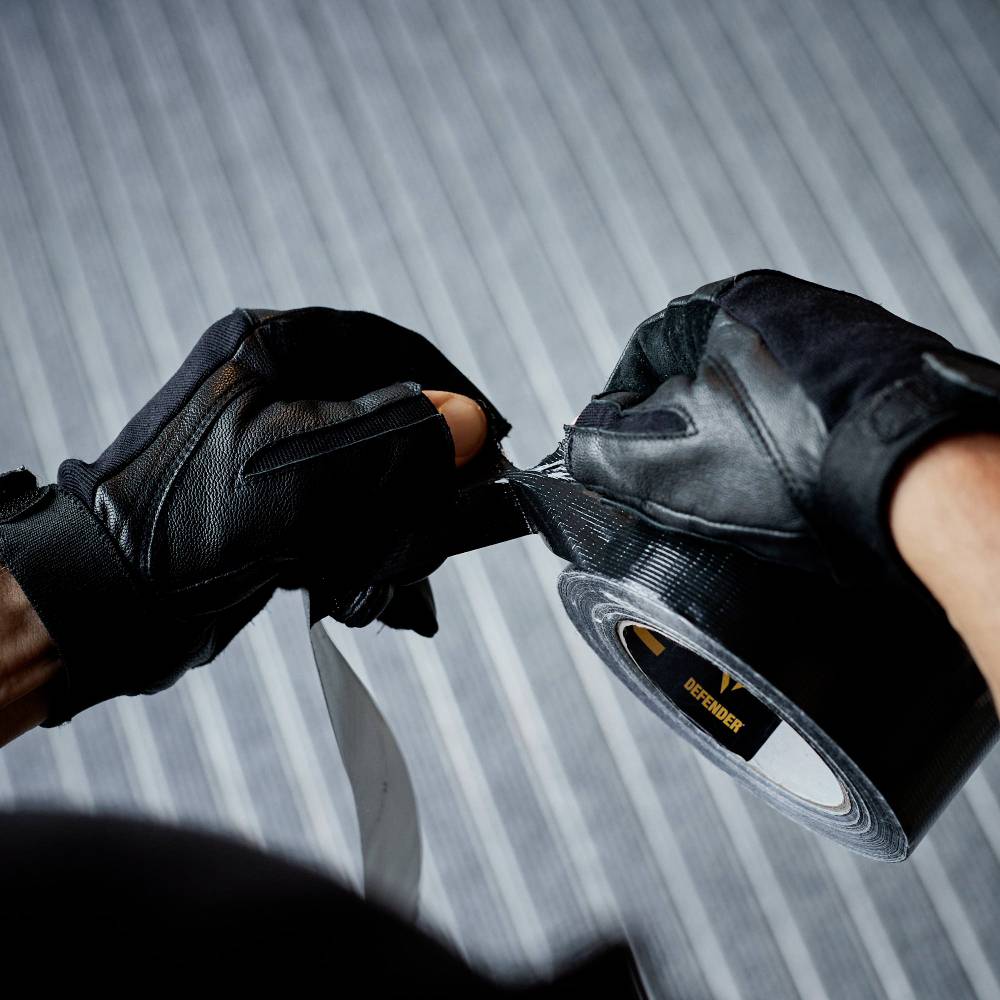 Nahaufnahme von verhandschuhten Händen, die ein Stück schwarzes Klebeband abreißen. Streifenmuster auf dem Boden im Hintergrund.