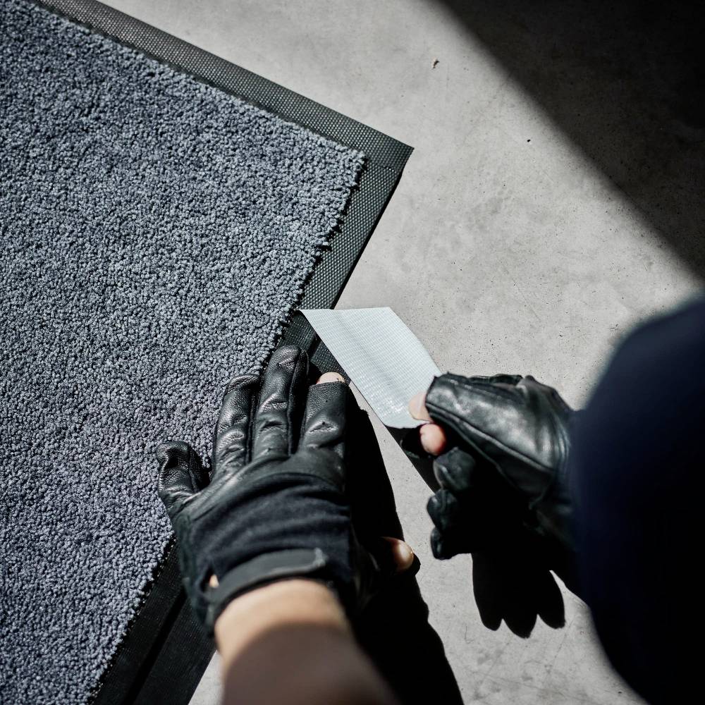 Person mit Handschuhen schneidet Teppichboden mit einem Messer. Der Teppich liegt auf grauem Boden, das Licht fällt von oben.