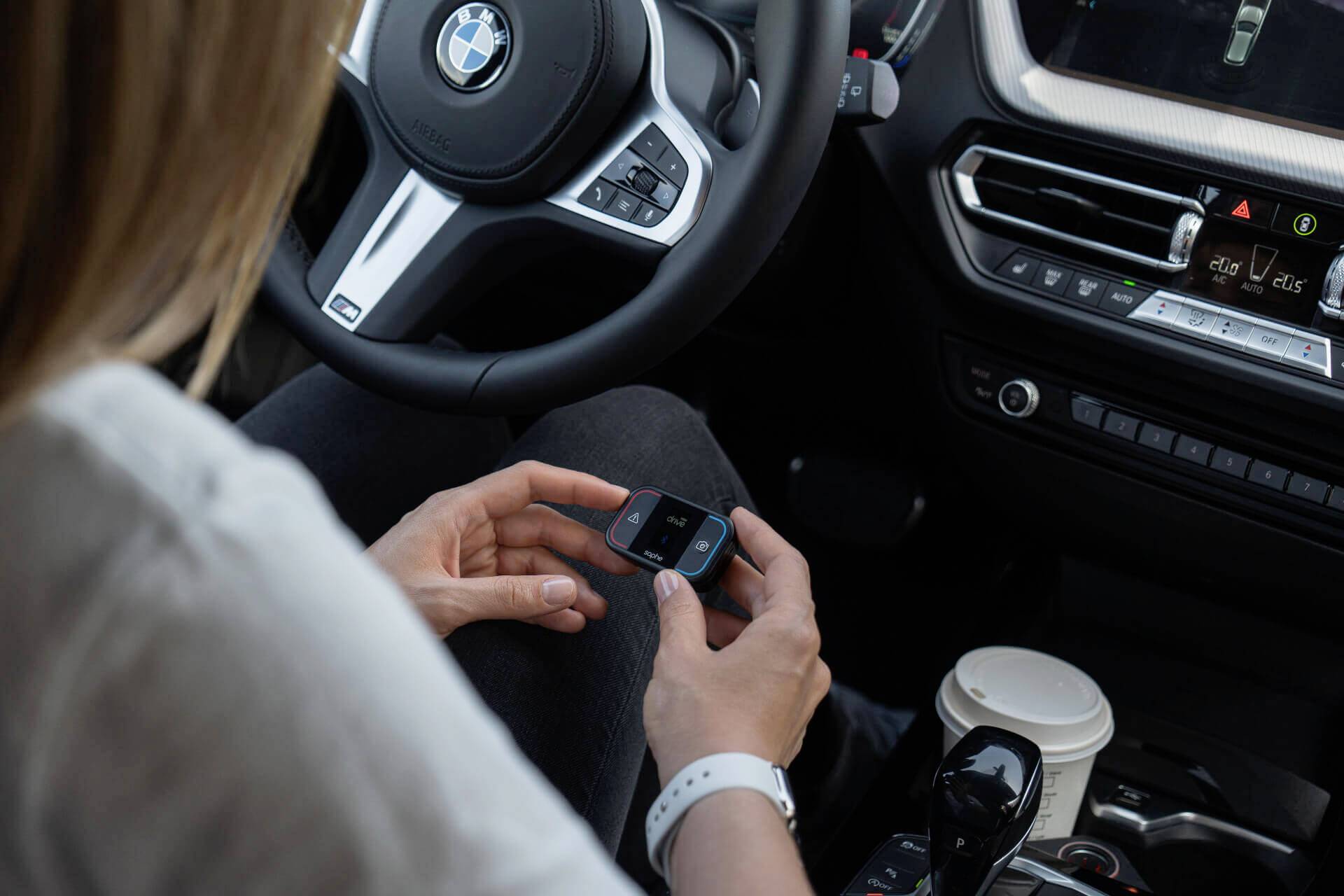 Eine Person sitzt im Auto, hält ein elektronisches Gerät in der Hand. Ein BMW-Logo ist auf dem Lenkrad sichtbar.