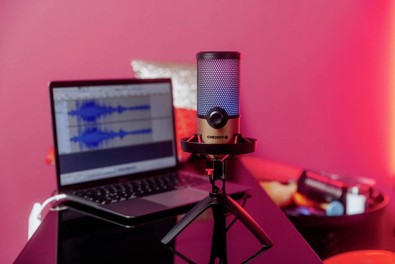 Ein Tischmikrofon steht vor einem aufgeklappten Laptop, der Audiospuren auf dem Bildschirm zeigt. Im Hintergrund ist eine rote Wand.