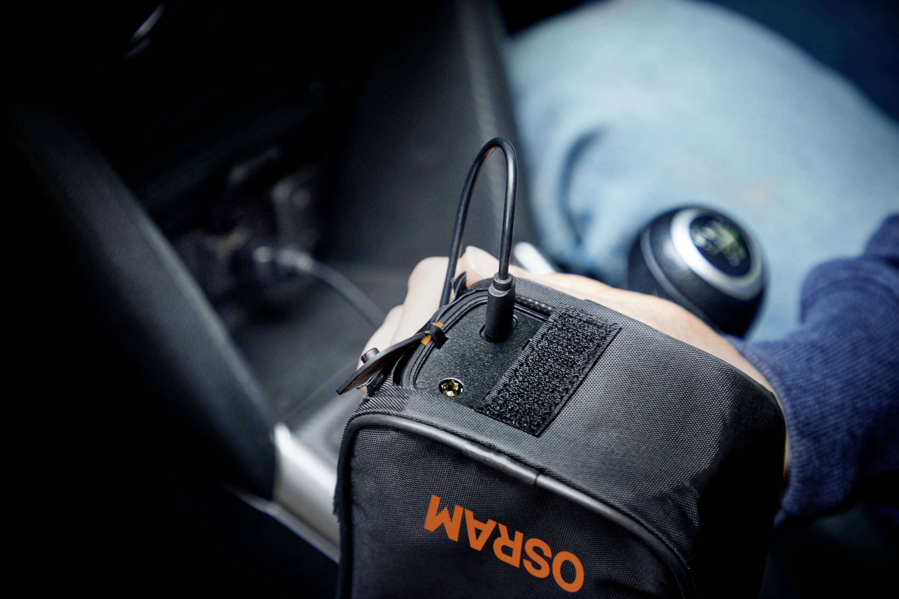 Eine Person im Auto lädt ein Gerät in einer Tasche mit 'OSRAM'-Aufschrift über eine Steckverbindung auf. Im Hintergrund ist der Schaltknauf sichtbar.