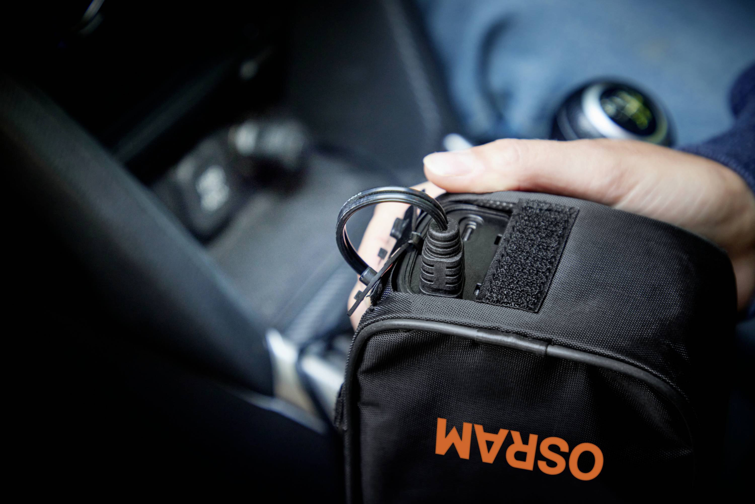 Eine Person hält eine schwarze Tasche mit der Aufschrift 'OSRAM', in die ein Ladekabel eingesteckt wird, im Inneren eines Fahrzeugs.