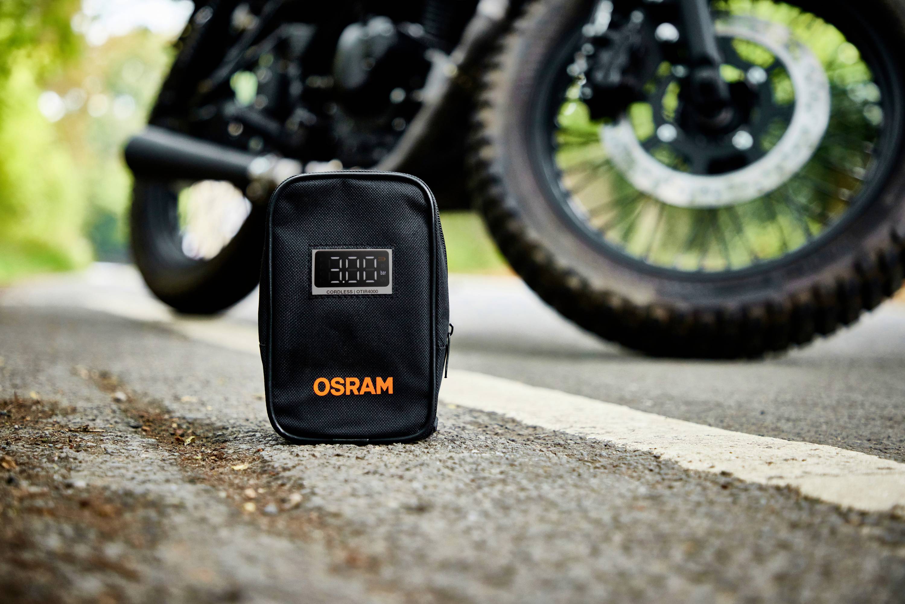 Eine schwarze Tasche mit der Aufschrift 'OSRAM' steht auf einer Straße, im Hintergrund ist ein Teil eines Motorrads zu sehen.