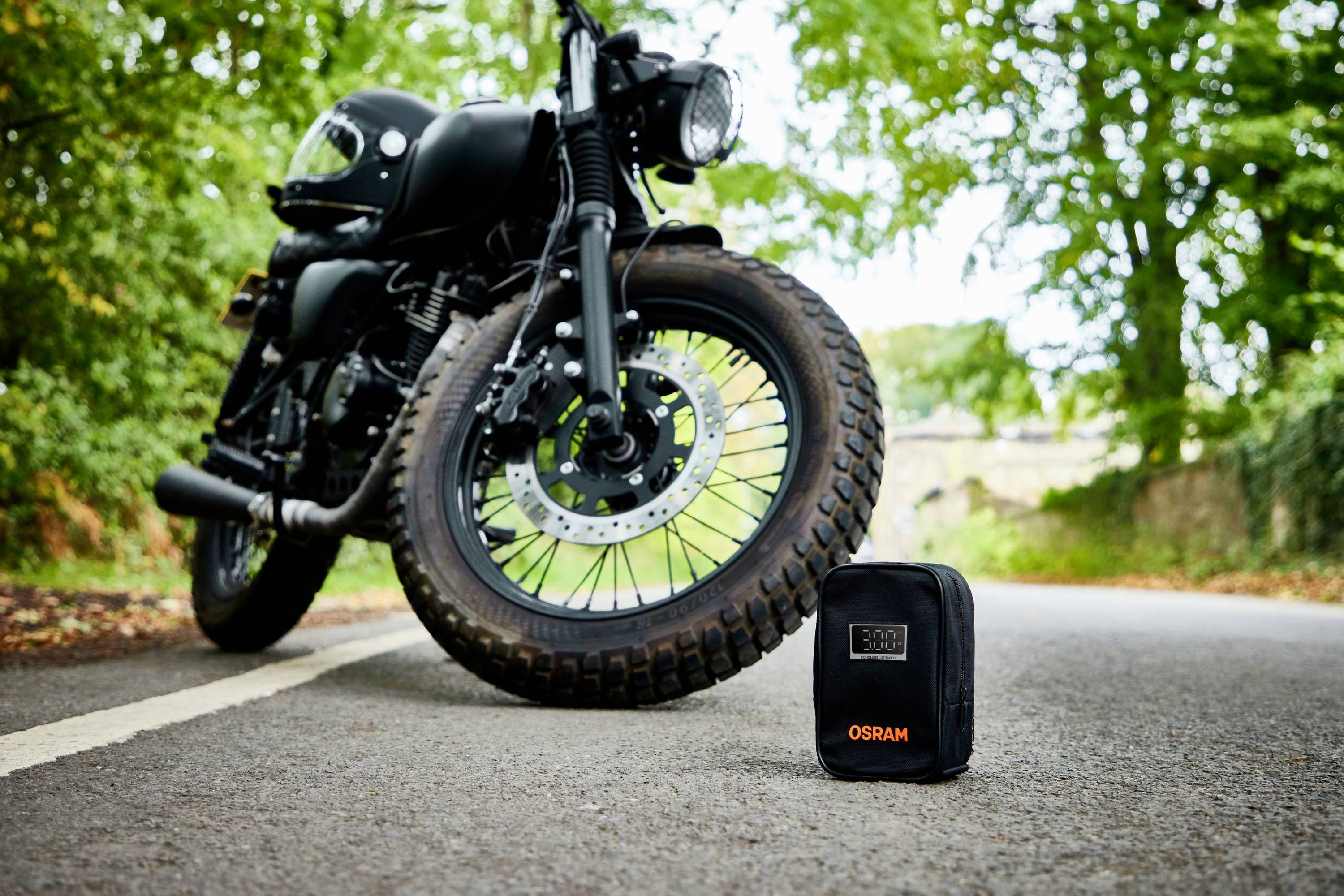 Schwarzes Motorrad steht auf einer Straße, flankiert von Bäumen. Im Vordergrund eine schwarze Tasche mit orange 'OSRAM'-Logo.