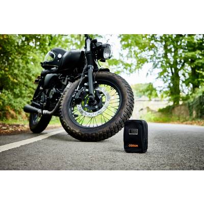 Schwarzes Motorrad steht auf einer Straße, flankiert von Bäumen. Im Vordergrund eine schwarze Tasche mit orange 'OSRAM'-Logo.