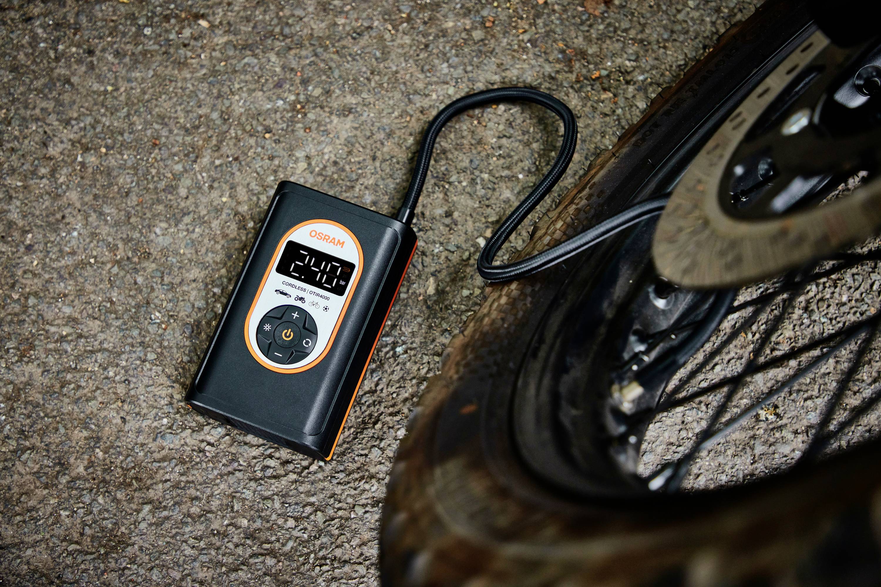 Ein tragbarer Luftkompressor liegt auf Asphalt neben einem Fahrradreifen, das Display zeigt '2.4' an.