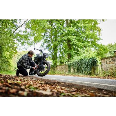 Ein Mann kniet neben einem Motorrad am Straßenrand und überprüft den Reifendruck. Im Hintergrund sind Bäume und eine Steinmauer zu sehen.