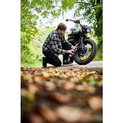 Eine Person kniet auf einer Straße und prüft den Reifendruck eines Motorrads im Grünen. Herbstblätter sind im Vordergrund sichtbar.