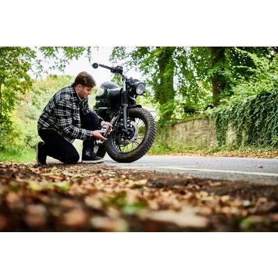 Ein Mann kniet auf einer Landstraße und überprüft den Reifendruck eines Motorrads mit einem Messgerät an einem bewaldeten Ort.