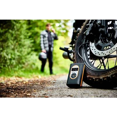 Ein Reifendruckmessgerät liegt neben einem Motorrad auf einem Waldweg. Im Hintergrund geht eine Person unscharf mit Helm in der Hand.