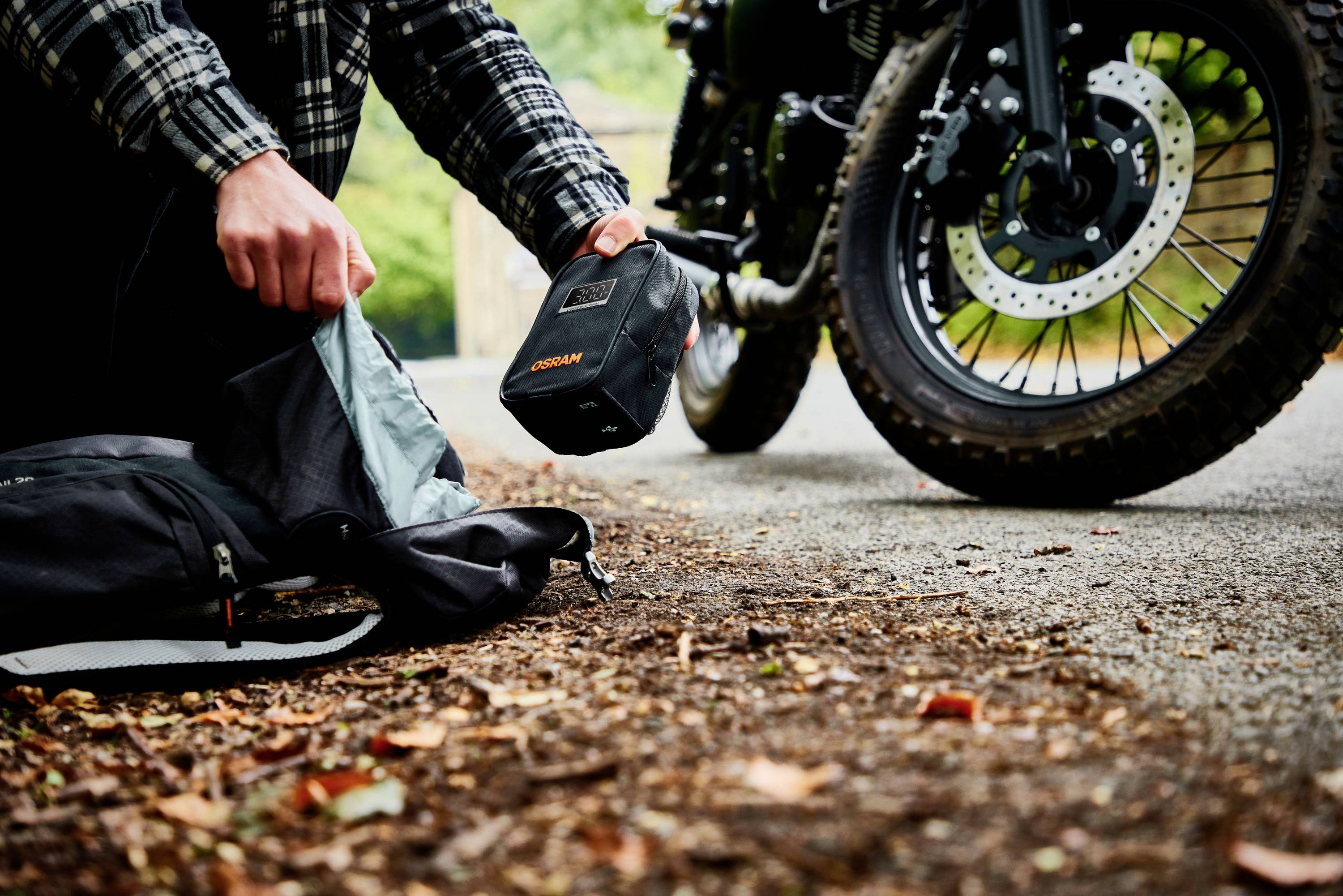 Eine Person packt Rucksack neben einem Motorrad aus, das auf einem Waldweg steht. Fokus auf Hände und Motorradreifen.
