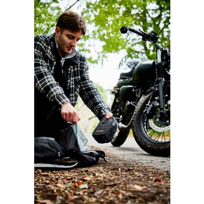 Ein Mann kniet neben einer offenen Tasche auf einem Waldweg und hält eine kleine Box. Im Hintergrund steht ein Motorrad.