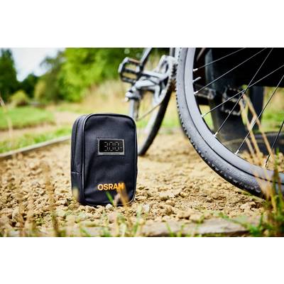 Eine schwarze Tasche mit 'OSRAM'-Aufdruck steht auf einem sandigen Weg neben einem Fahrradreifen, im Hintergrund sind Bäume sichtbar.