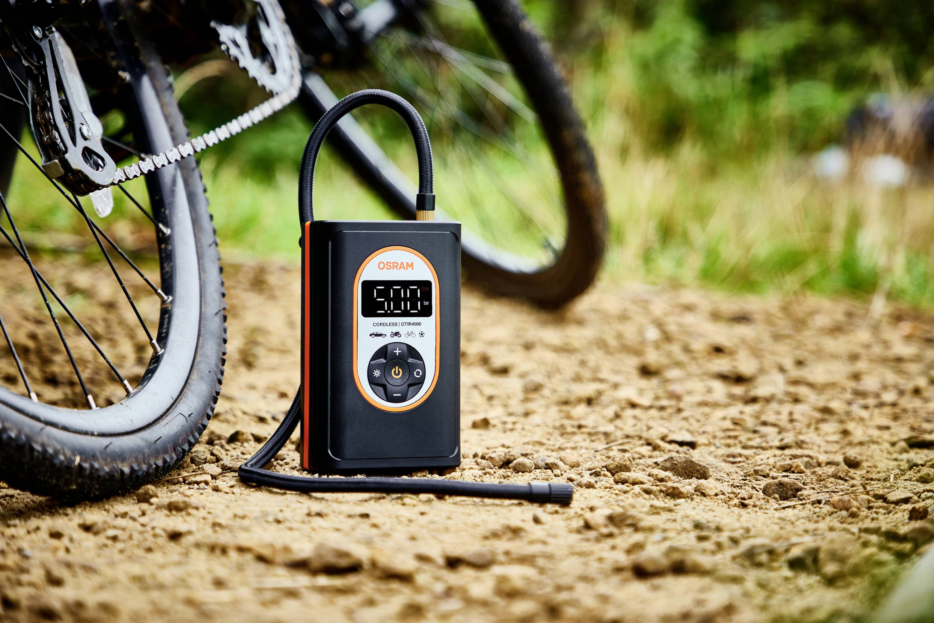 Ein elektrischer Luftkompressor steht auf einem unbefestigten Weg neben einem Fahrrad, bereit zum Aufpumpen der Reifen.
