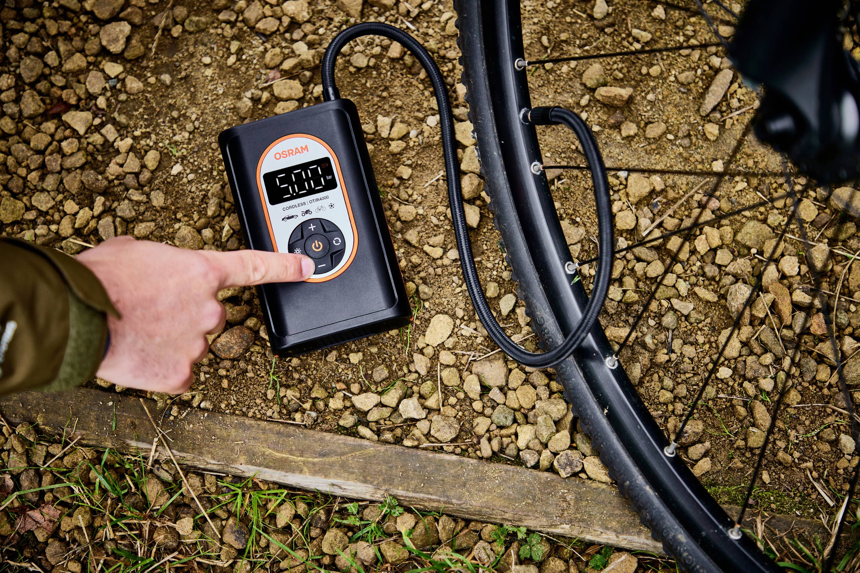 Eine Hand bedient einen digitalen Luftkompressor, der einen Fahrradreifen auf einem steinigen Untergrund aufpumpt.
