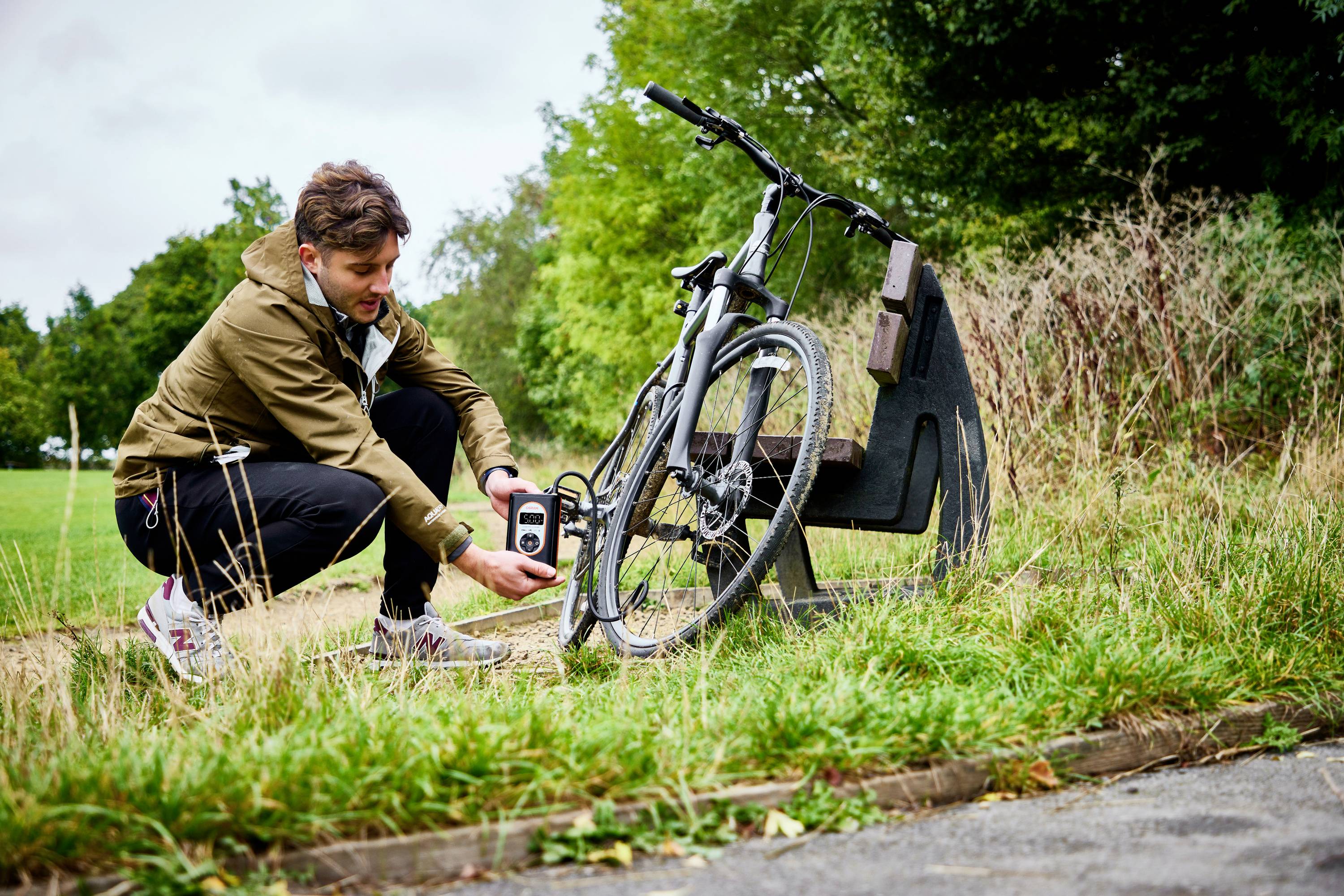 Ein Mann schließt ein Fahrrad an einem Fahrradständer im Park mit einem Schloss ab. Im Hintergrund sind Bäume und Rasen zu sehen.