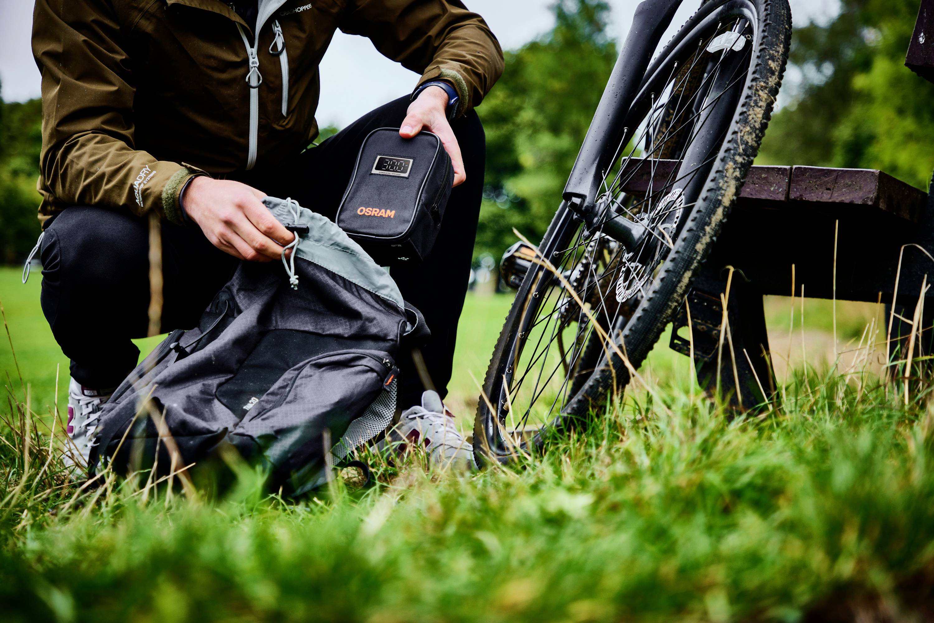 Eine Person packt an einem Fahrrad eine kleine Tasche in einen Rucksack ein. Im Hintergrund ist grüne Landschaft zu sehen.