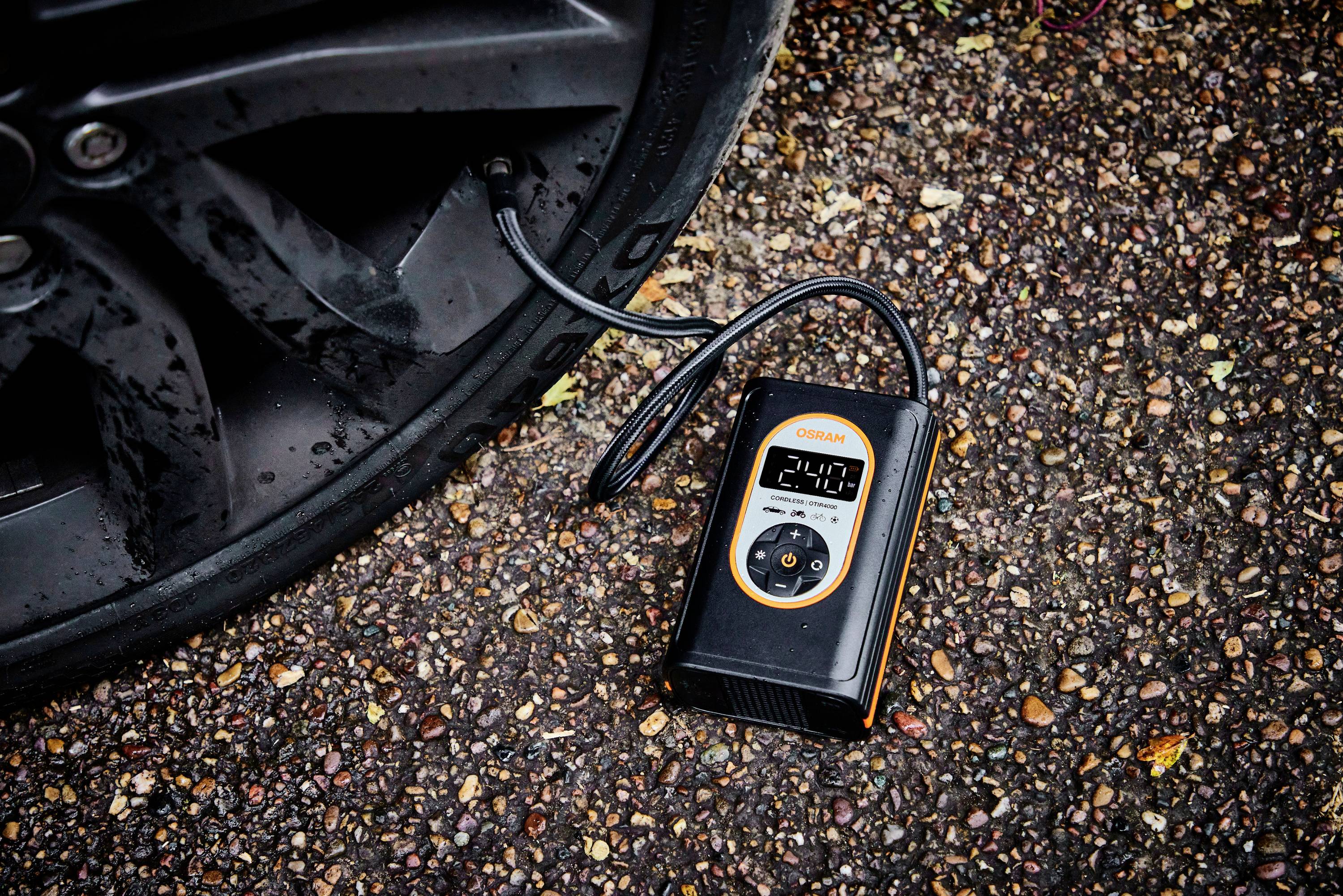 Ein digitaler Reifenfüller überprüft den Druck eines Autoreifens auf einer Kieseloberfläche. Das Display zeigt '2,4 bar' an.