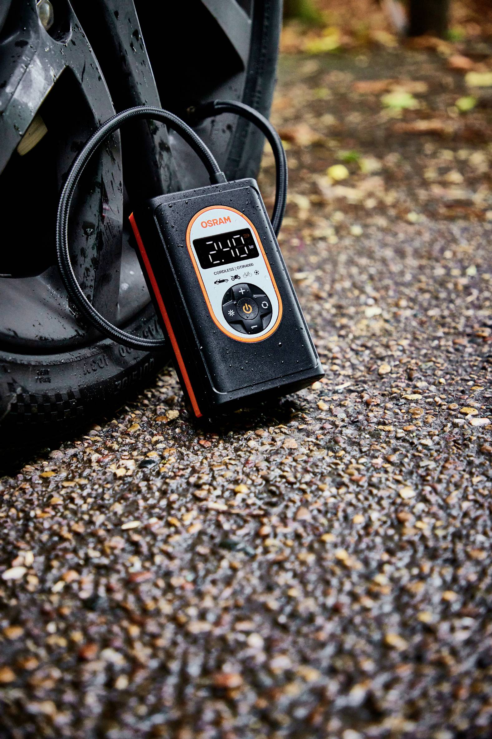 Tragbarer Reifenluftkompressor neben einem Autoreifen auf regennassem Asphalt; Display zeigt Reifendruck von 2,4 bar.