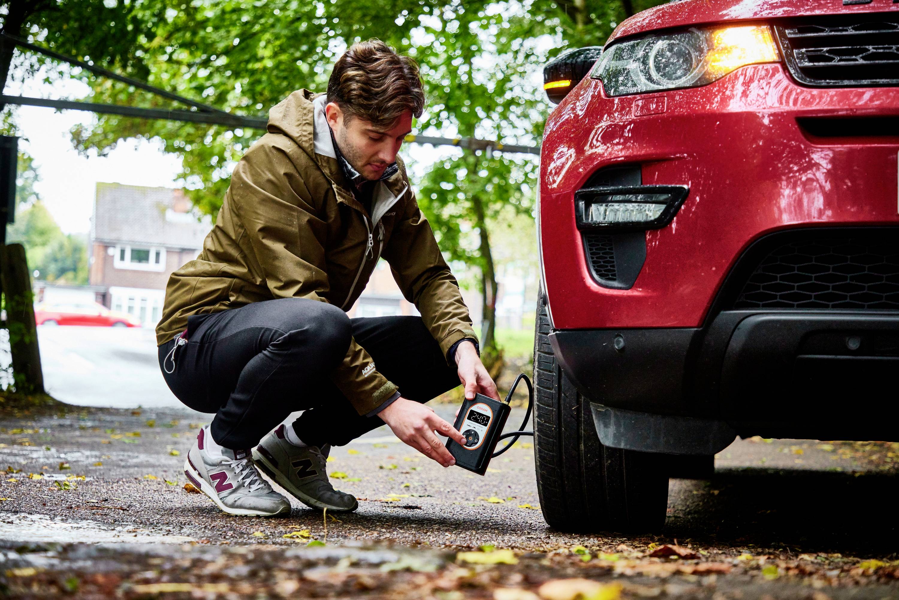Ein Mann überprüft den Reifendruck eines roten Autos mit einem Reifendruckprüfer auf einem bewaldeten Parkplatz.