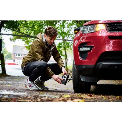 Ein Mann überprüft den Reifendruck eines roten Autos mit einem Reifendruckprüfer auf einem bewaldeten Parkplatz.