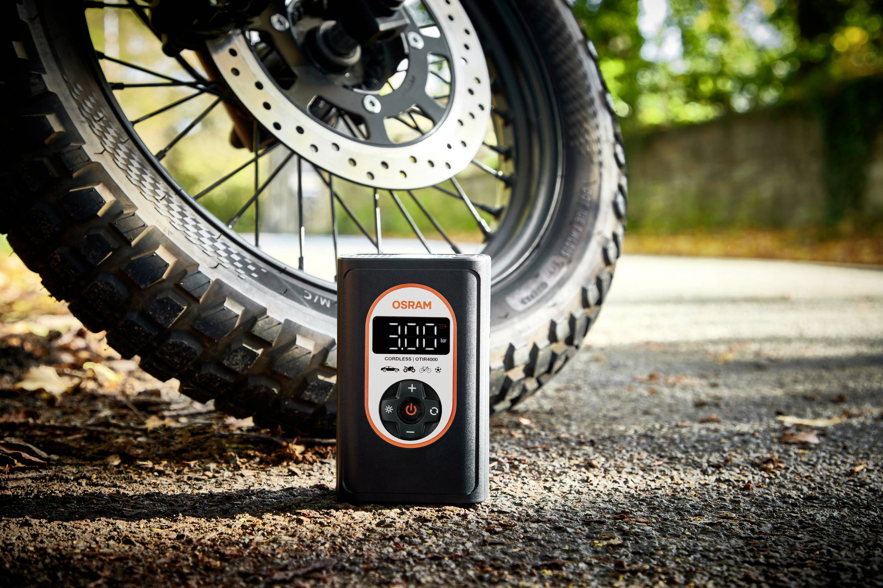 Ein Reifenfülldruckmesser zeigt 30,0 an und steht vor einem Motorradreifen auf einer sonnigen Straße.