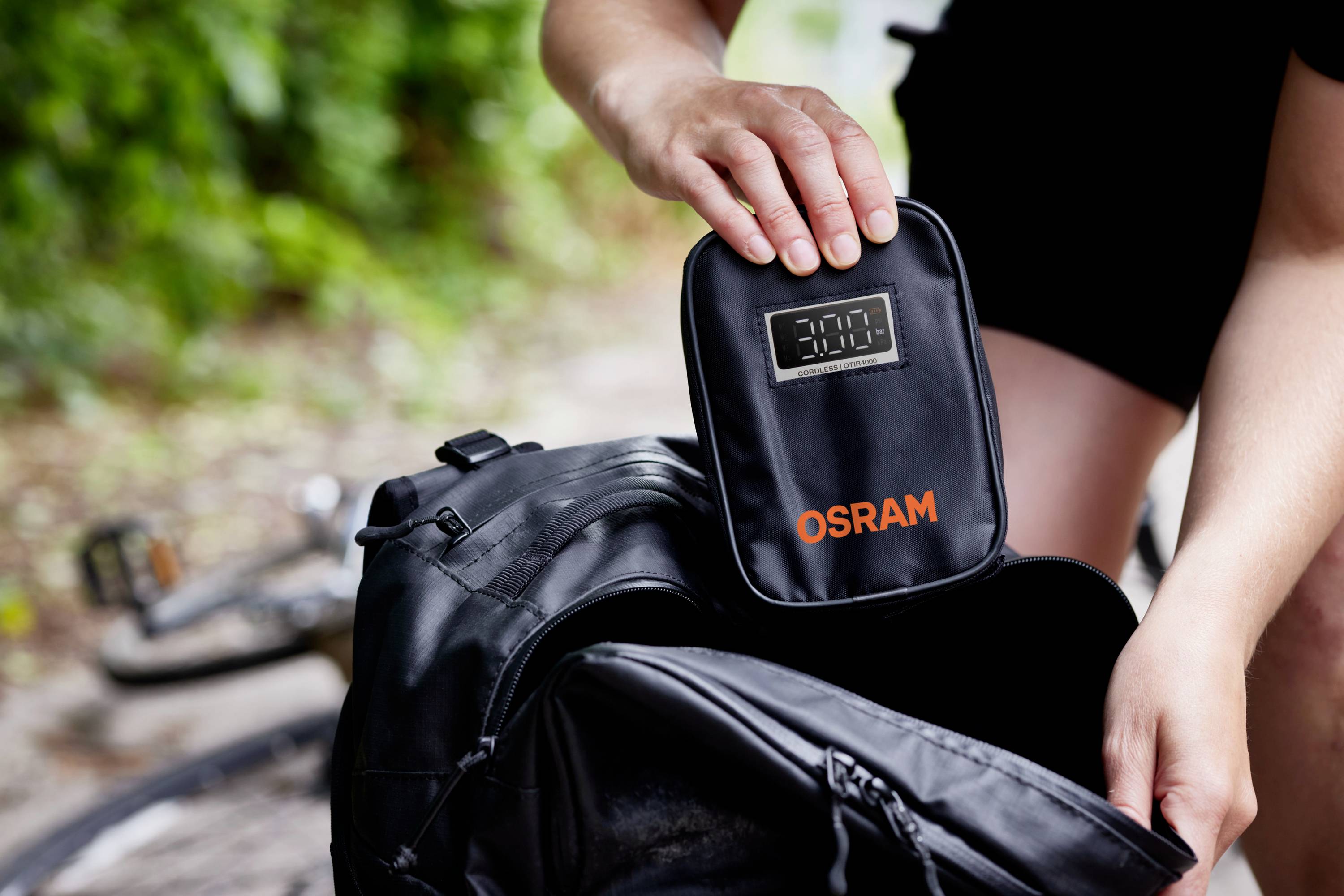 Eine Person packt eine kleine schwarze Tasche mit der Aufschrift 'OSRAM' in einen Rucksack. Im Hintergrund ist ein Fahrrad sichtbar.