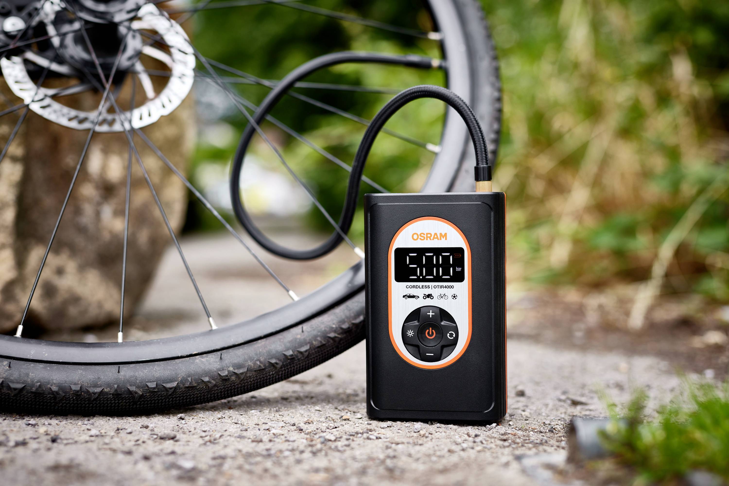 Ein digitales Manometer pumpt Fahrradreifen auf. Der Bildschirm zeigt '5,00'. Im Hintergrund sind Fahrrad und Büsche zu sehen.