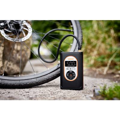 Ein digitales Manometer pumpt Fahrradreifen auf. Der Bildschirm zeigt '5,00'. Im Hintergrund sind Fahrrad und Büsche zu sehen.