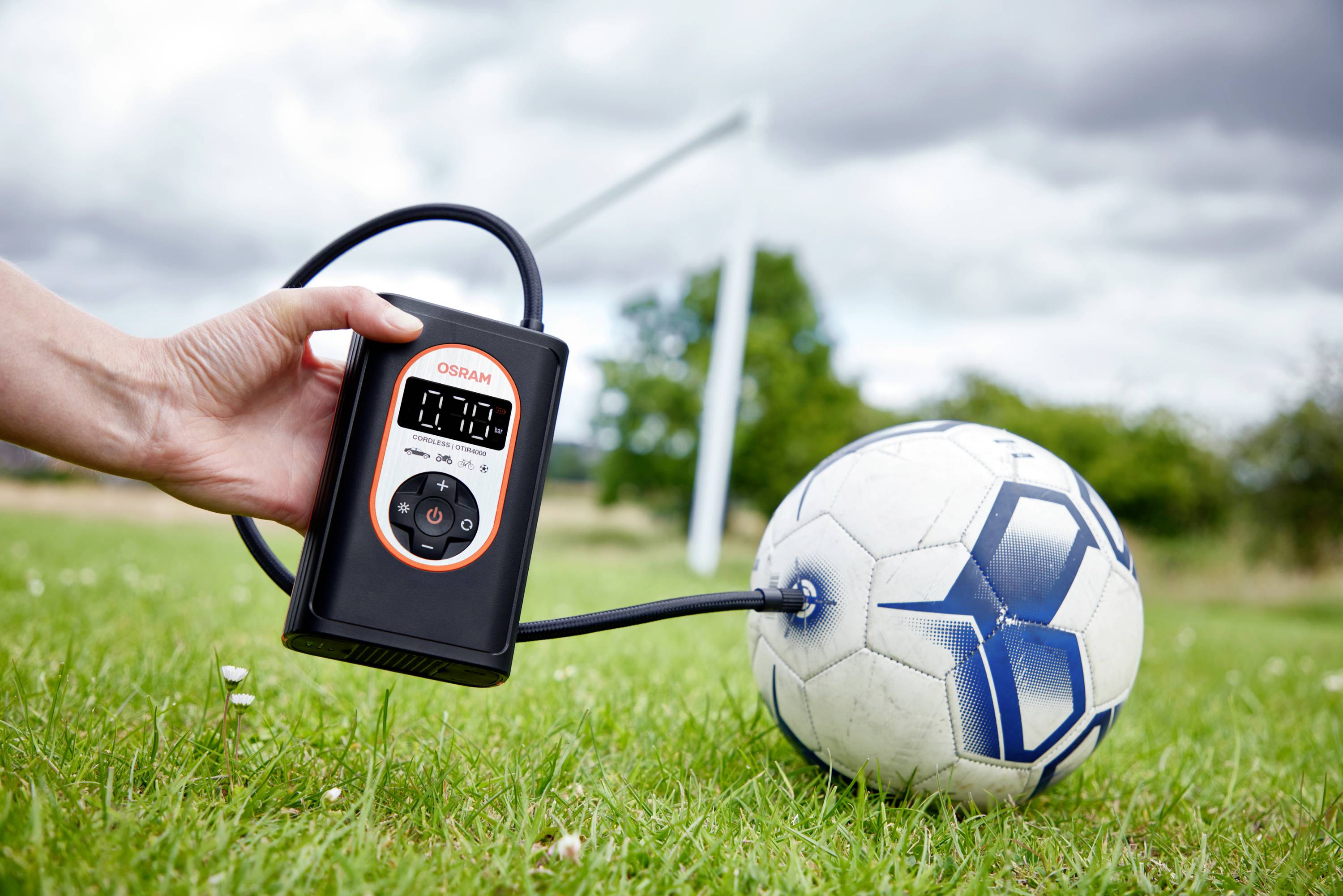 Person pumpt Fußball mit einem tragbaren elektrischen Kompressor auf. Der Ball liegt auf einer grünen Wiese, ein Tor ist im Hintergrund.