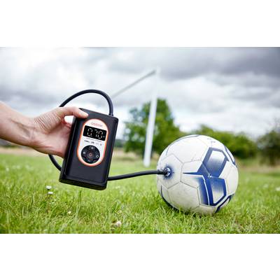 Person pumpt Fußball mit einem tragbaren elektrischen Kompressor auf. Der Ball liegt auf einer grünen Wiese, ein Tor ist im Hintergrund.