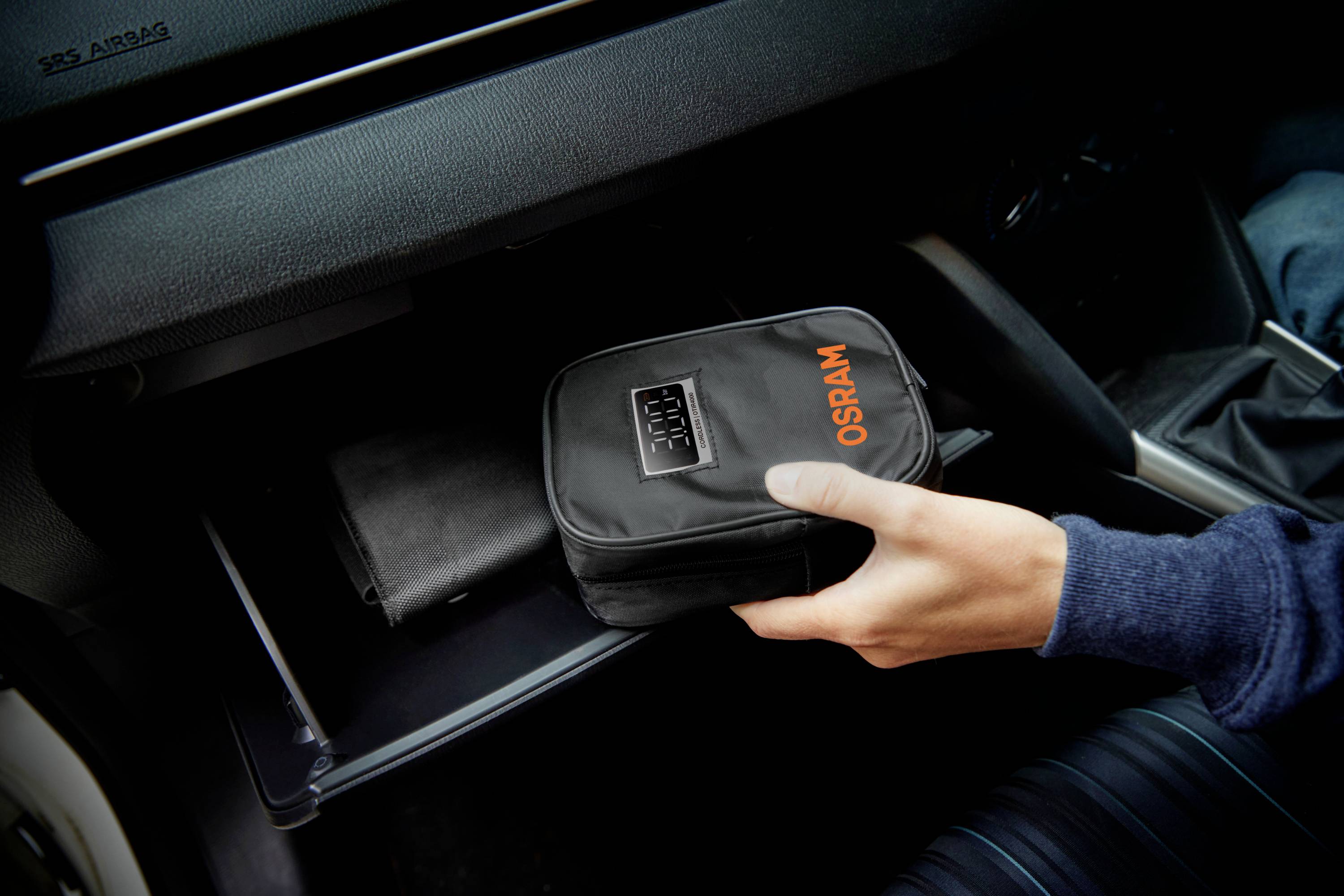 Hand legt eine schwarze Tasche mit 'OSRAM'-Logo in ein geöffnete Handschuhfach im Auto.