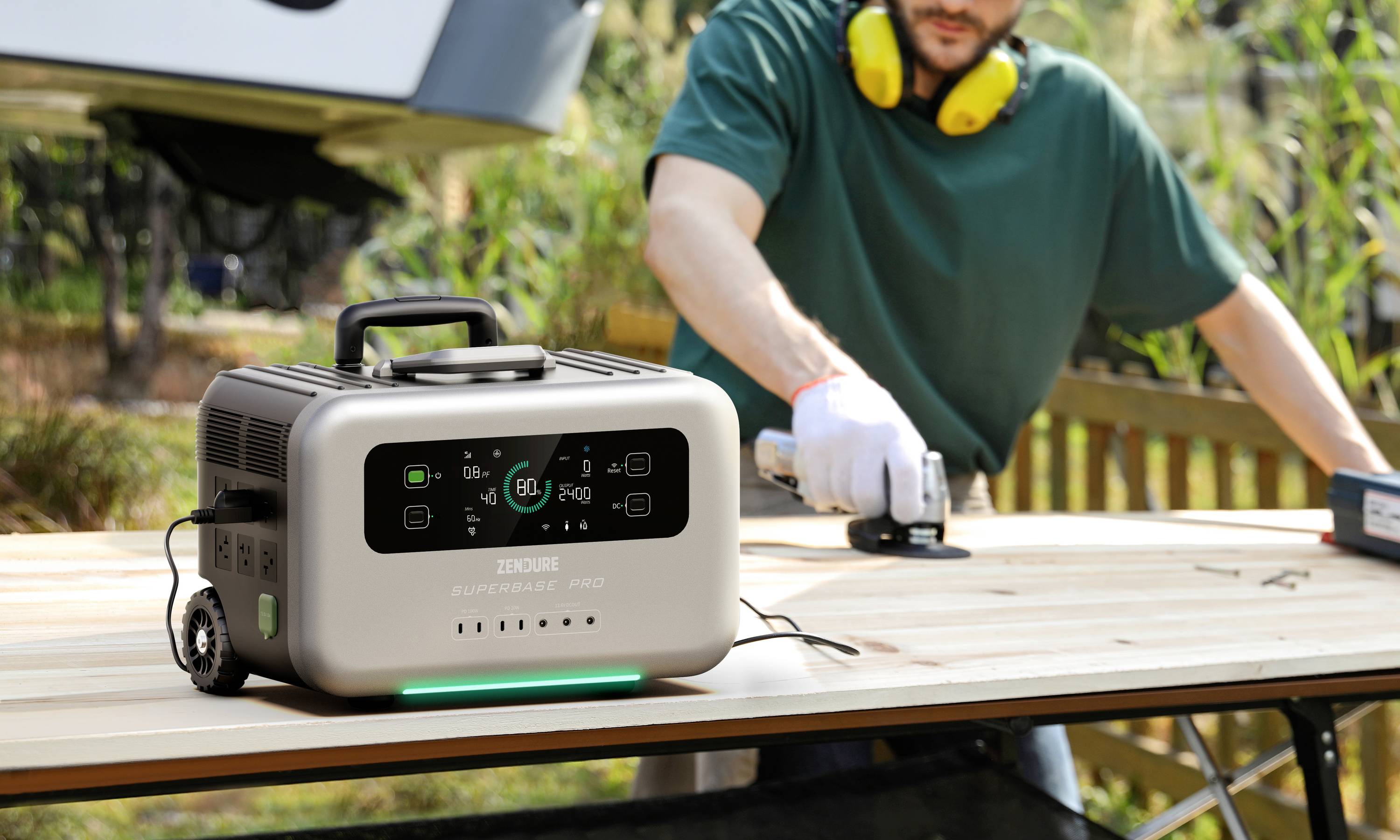 Ein Mann arbeitet an einem Holztisch im Freien mit einem Handwerkzeug, während ein tragbarer Stromgenerator in der Nähe steht.