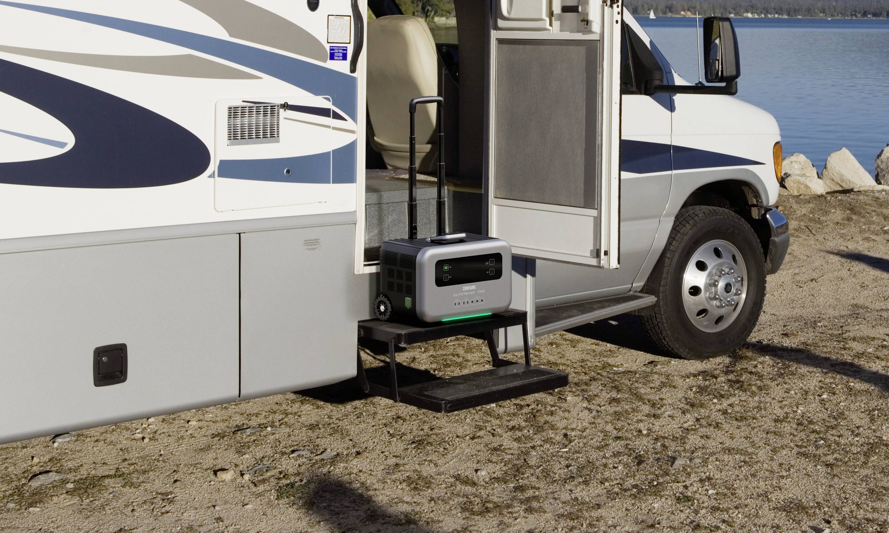 Ein tragbarer Energiespeicher steht auf einer Stufe vor einem Wohnmobil, das an einem See geparkt ist. Im Hintergrund sind Wasser und Steine zu sehen.