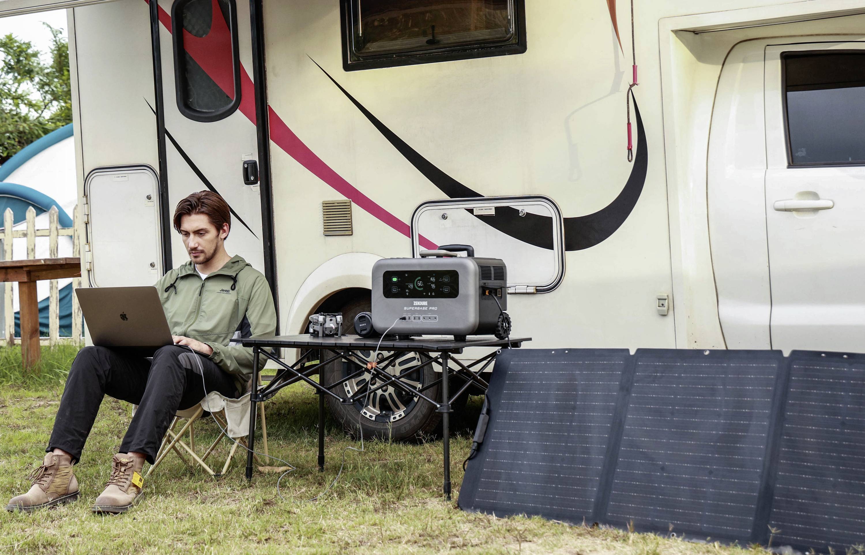 Ein Mann sitzt auf einem Campingstuhl neben einem Wohnmobil und arbeitet an einem Laptop. Neben ihm steht ein tragbarer Generator, der an Solarpanels angeschlossen ist.