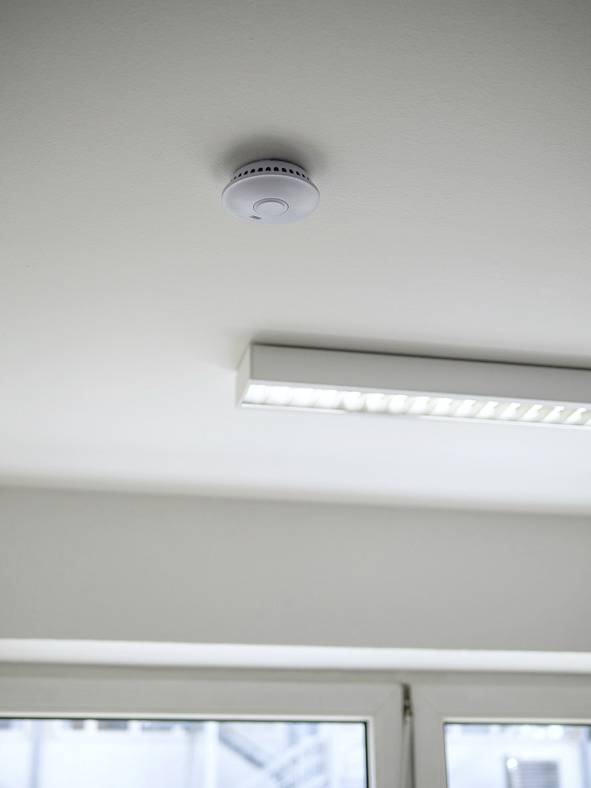 Ein Rauchmelder ist an einer weißen Zimmerdecke nahe einer Leuchtstofflampe angebracht, wichtige Sicherheitsvorrichtung im Raum.