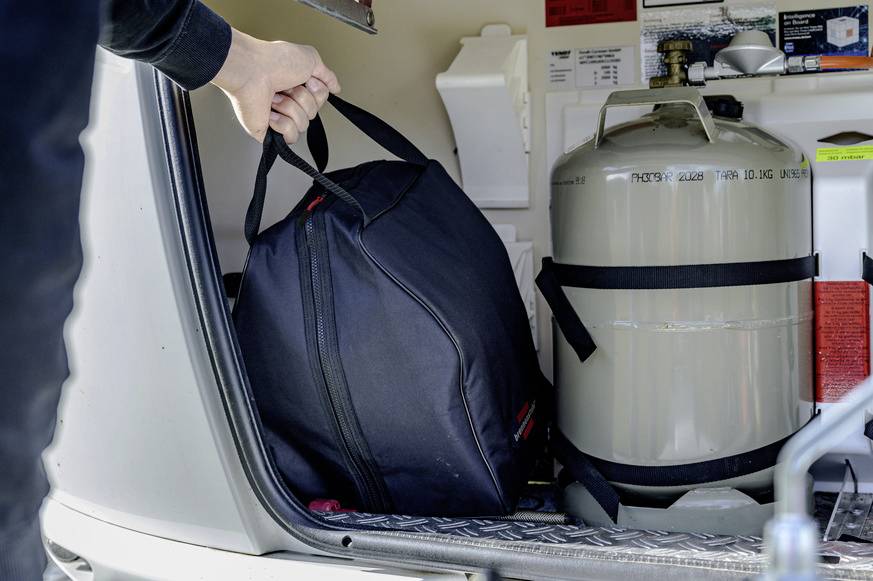 Eine Person legt eine schwarze Tasche in den Kofferraum eines Fahrzeugs neben einen großen Metalltank.