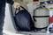 Eine Person legt eine schwarze Tasche in den Kofferraum eines Fahrzeugs neben einen großen Metalltank.