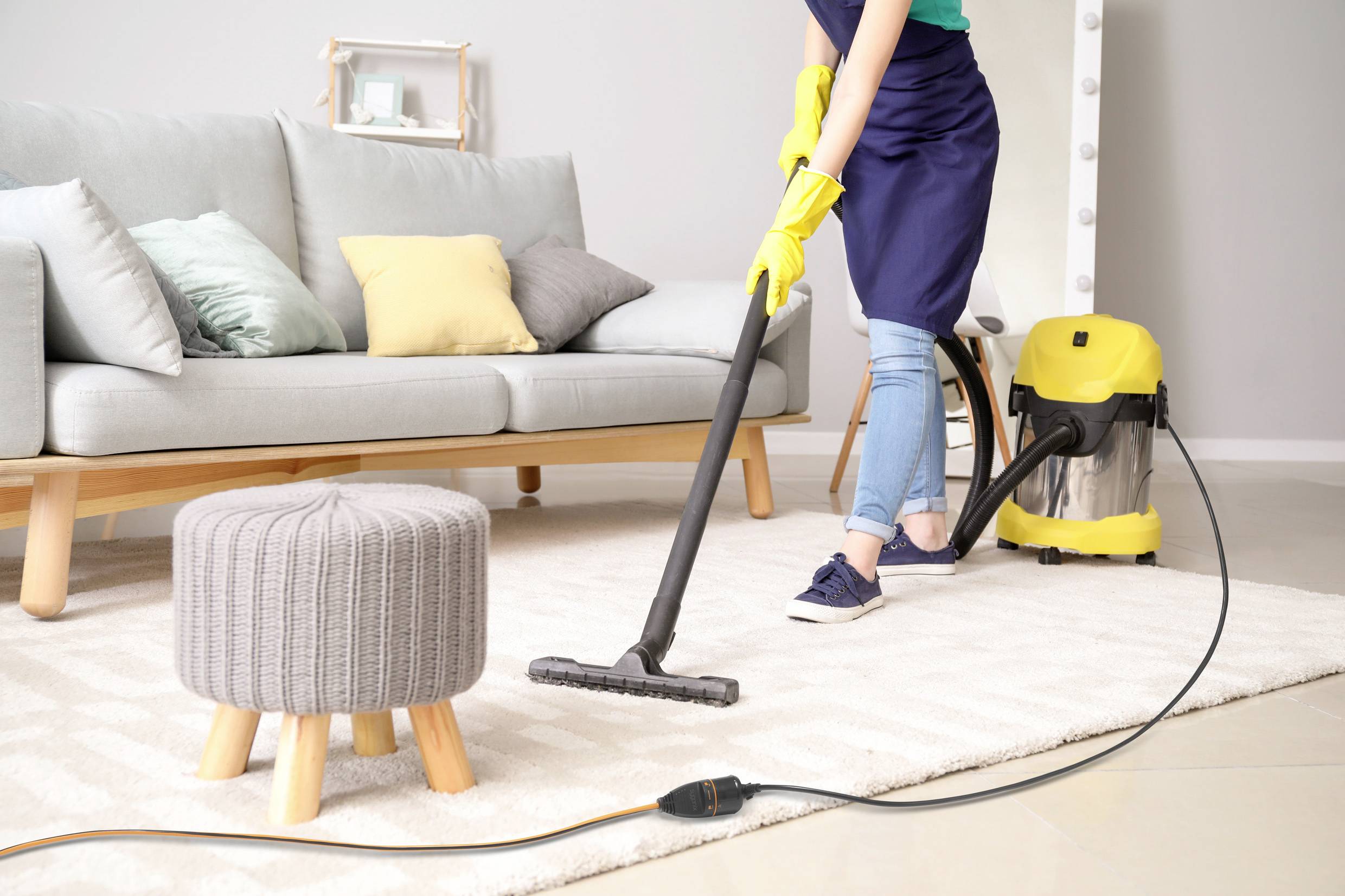 Person saugt mit Staubsauger einen hellen Teppich in einem Wohnzimmer. Im Hintergrund sind ein graues Sofa und bunte Kissen sichtbar.