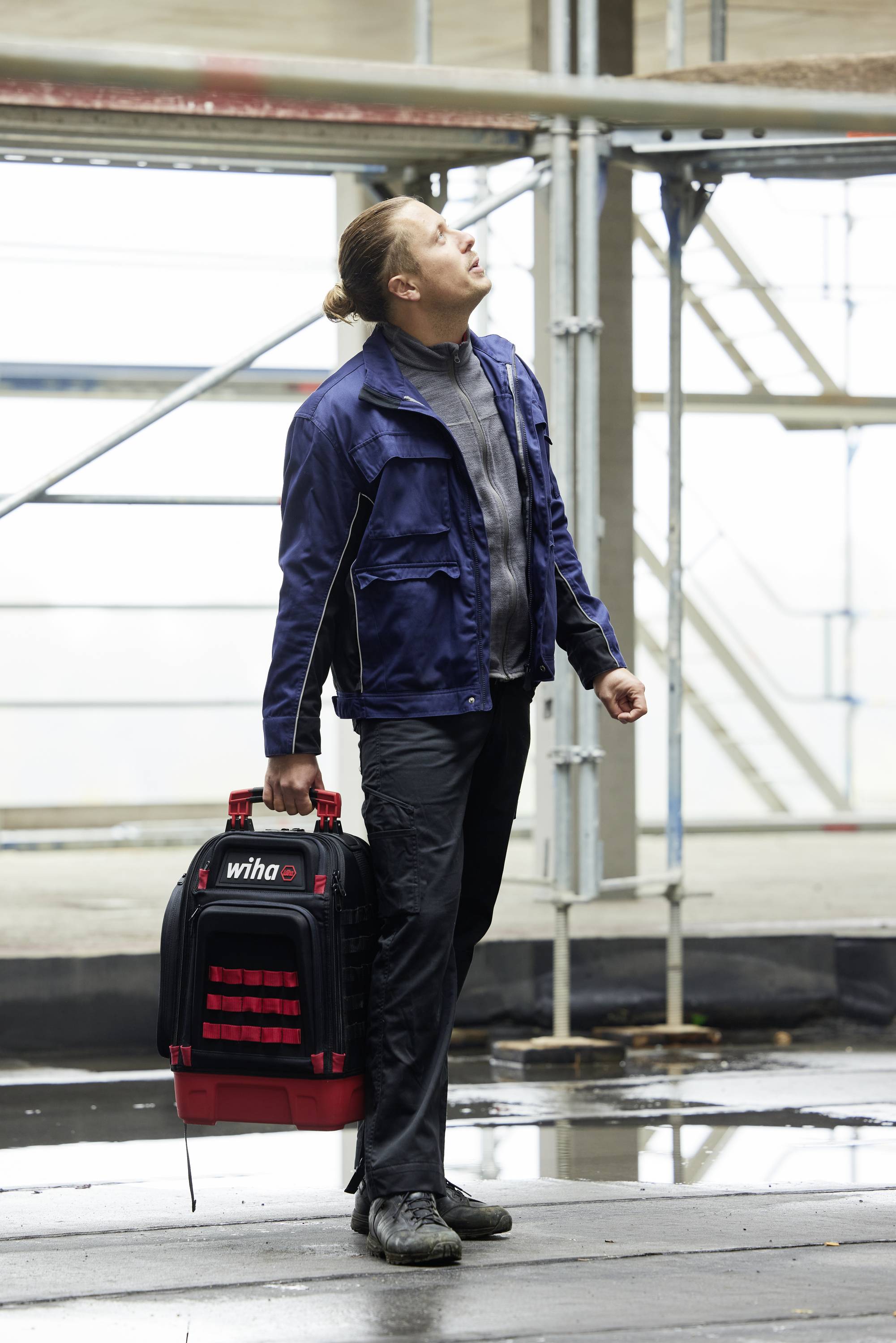 Person in Arbeitskleidung steht auf einer Baustelle und hält einen Werkzeugkasten. Sie schaut nach oben. Im Hintergrund ist ein Gerüst sichtbar.