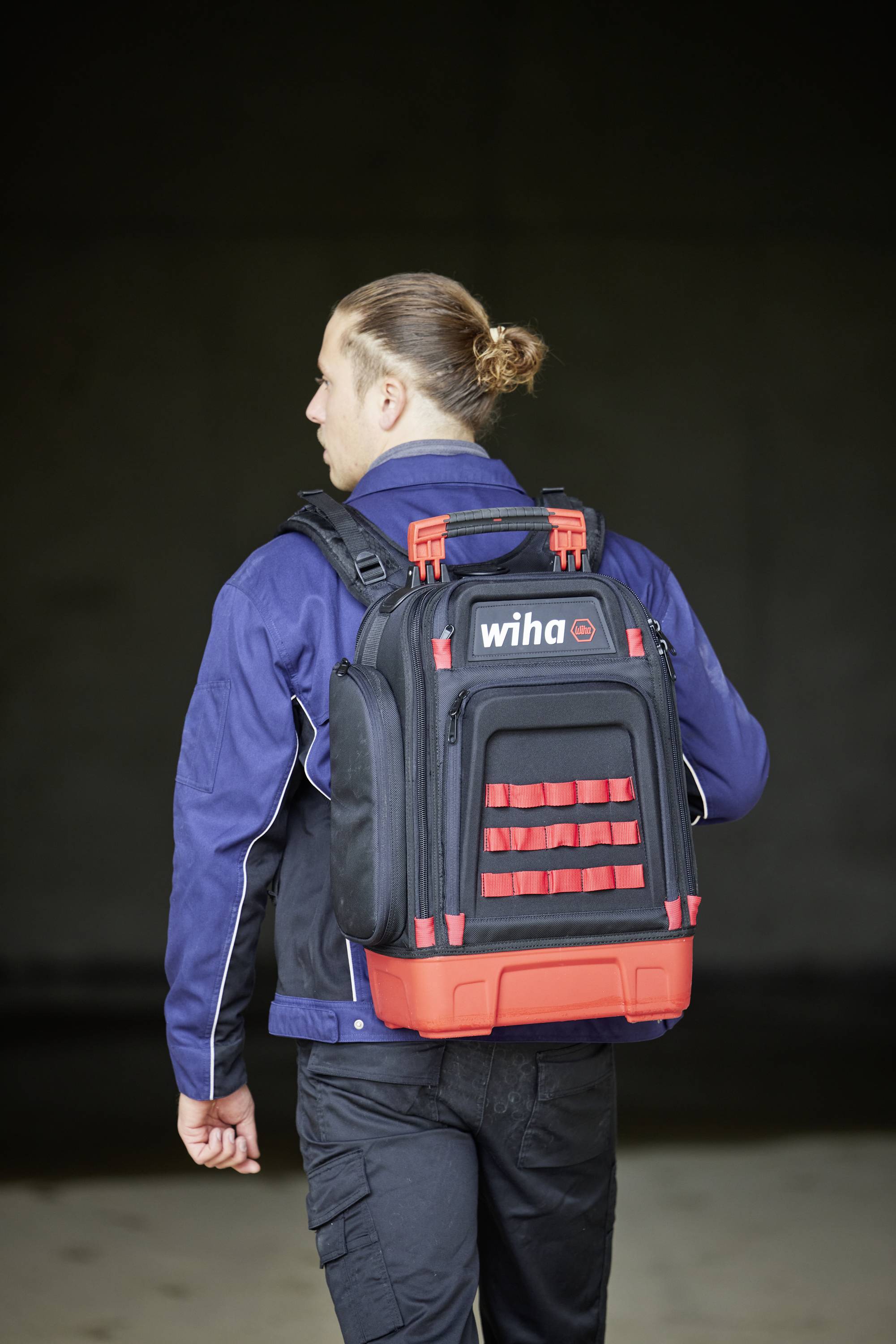 Eine Person trägt einen schwarzen und roten Rucksack der Marke 'Wiha'. Sie trägt eine blaue Jacke und schwarze Hosen.