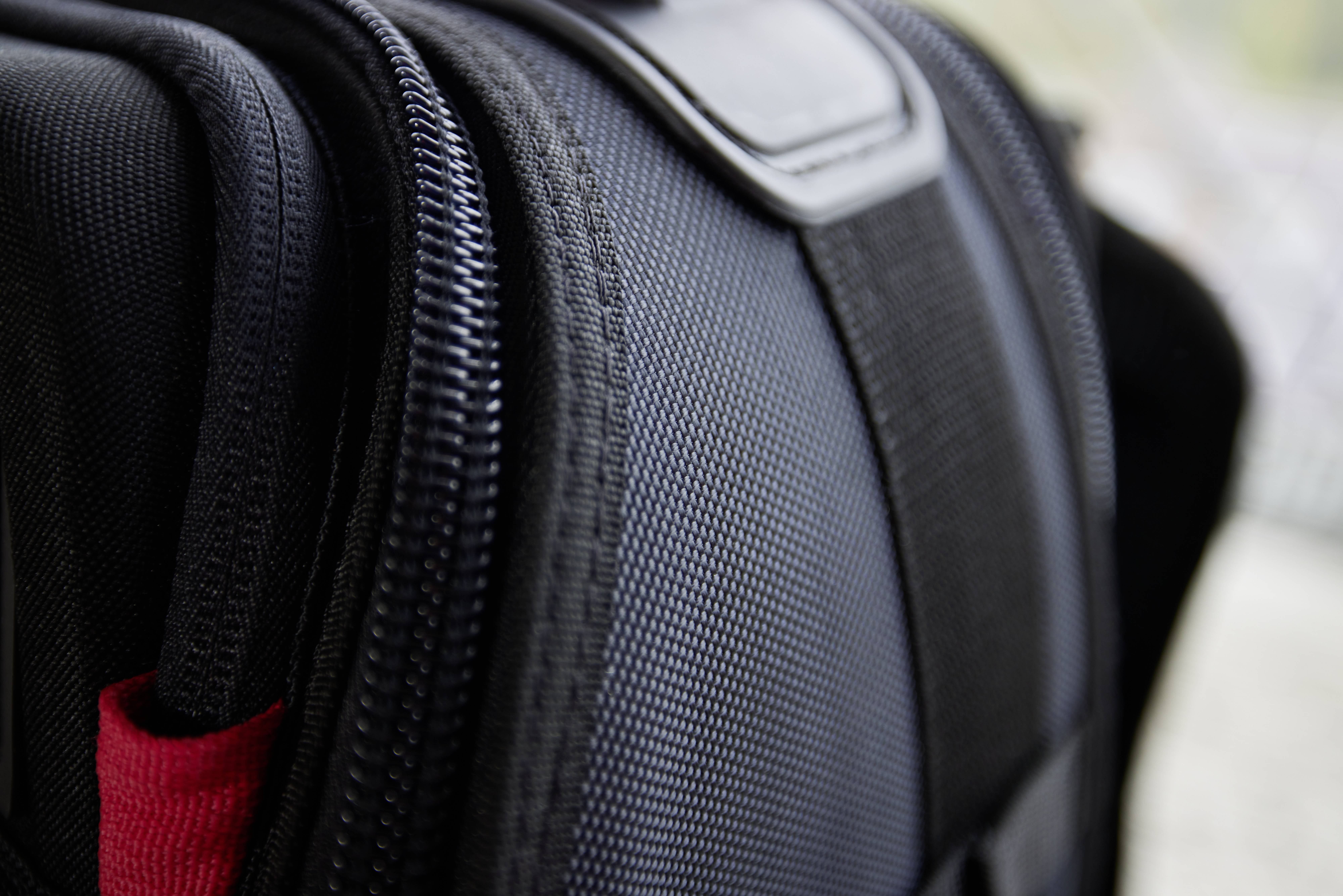 Nahaufnahme eines schwarzen Rucksacks mit sichtbaren Reißverschlüssen und einem roten Detail. Minimalistisches Design im Vordergrund.