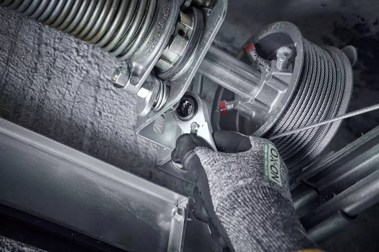 Hand in Schutzhandschuh dreht Schraube an einem metallischen Garagentormechanismus mit Feder und Seilzug. Mechanikerarbeit.