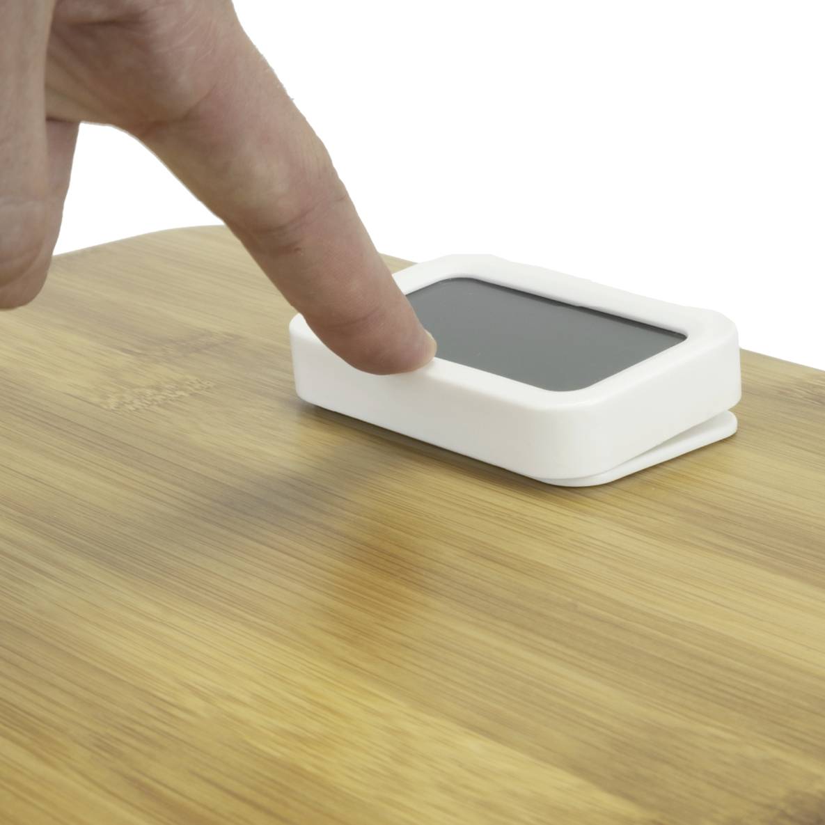 Eine Hand berührt einen kleinen, rechteckigen, weißen Sensor, der auf einem Holztisch liegt. Der Sensor ist glatt und modern gestaltet.