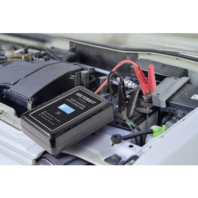 Ein Ladegerät ist an eine Autobatterie angeschlossen. Das Gerät zeigt einen Ladevorgang an. Im Hintergrund ist der Motorraum sichtbar.