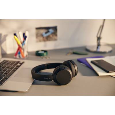 Ein Laptop und schwarze Kopfhörer liegen auf einem Schreibtisch. Daneben sind Stifte, ein Notizblock und eine Lampe zu sehen.